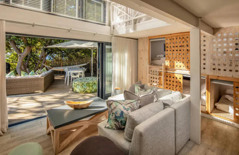 Spacious open-plan living room in seaside bungalow with large glass doors to ocean-view deck, plush sofa, wooden table.