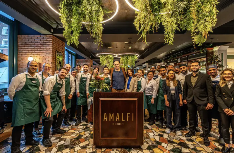 Group of Amalfi Ristorante staff in green aprons smiling in front of 'AMALFI' podium, stylish interior with hanging plants and tiled floor.