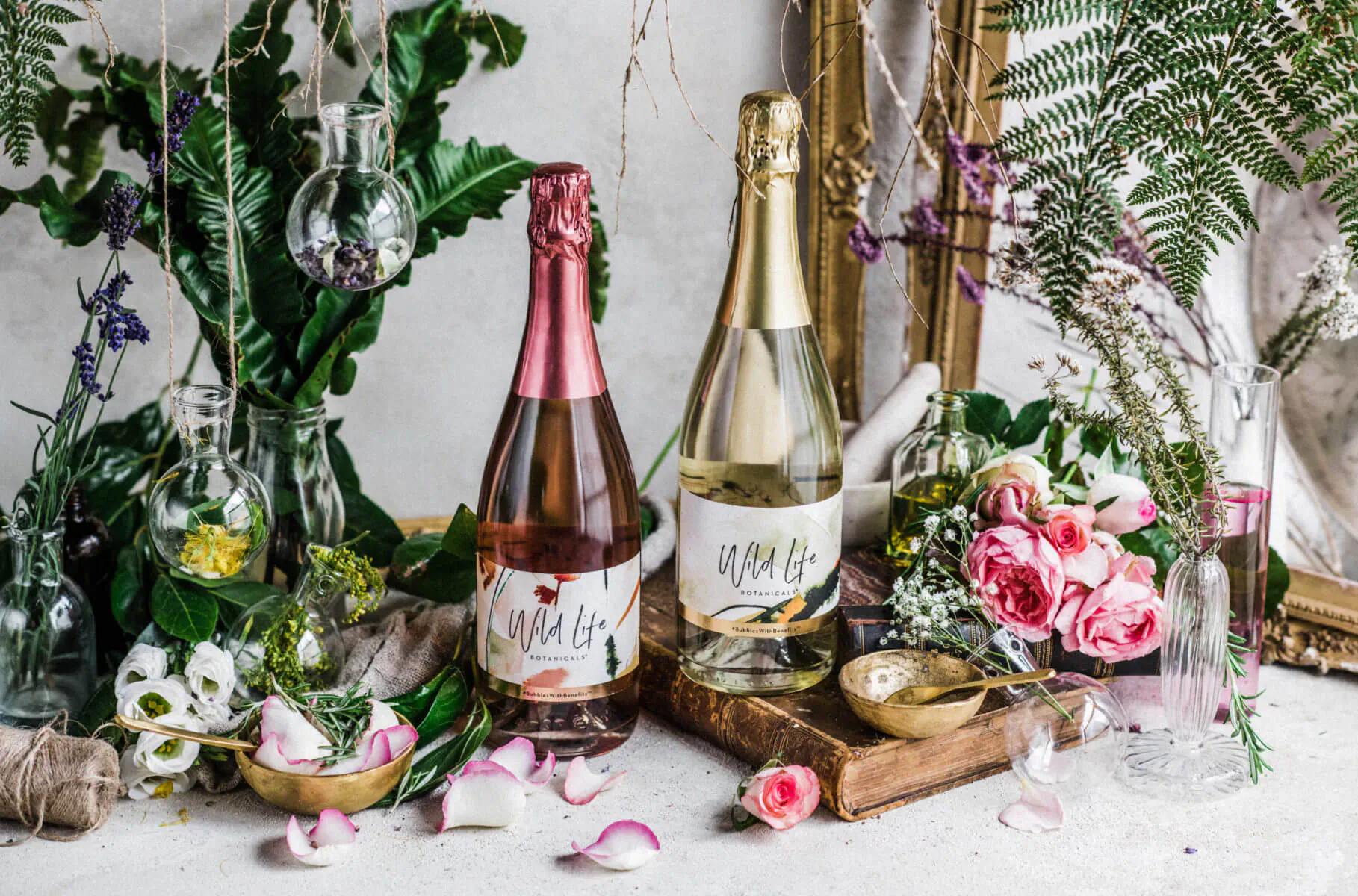 Two Wild Life Botanicals wine bottles, pink rosé and white, with ferns, pink roses, and gold spoon on wooden board