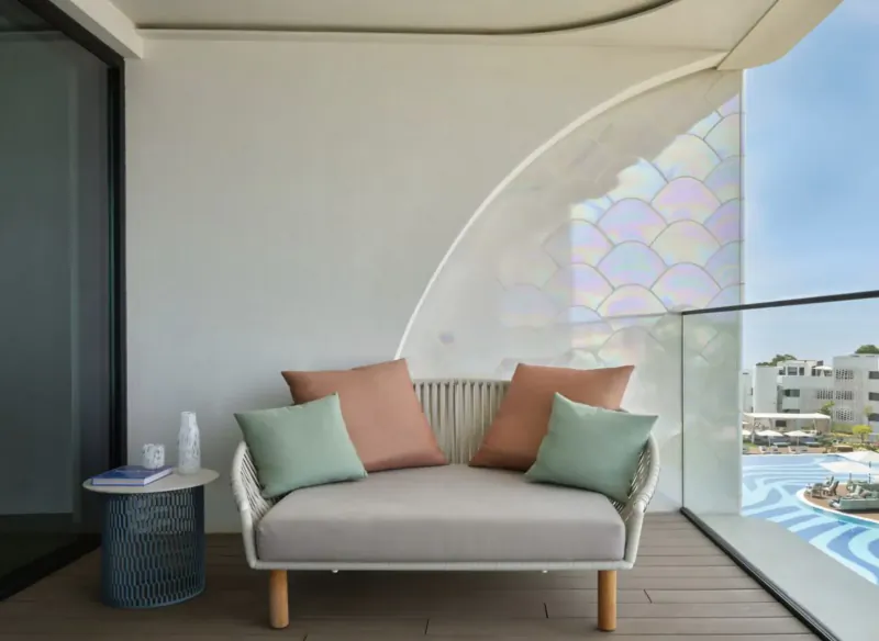 Cozy loveseat with orange and green cushions on Algarve hotel balcony overlooking pool, iridescent window accents.