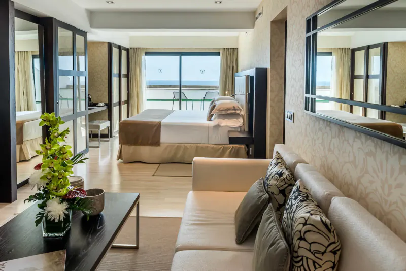 Luxurious Nixe Palace hotel suite with king bed, beige sofa, orchids, and sea view balcony.