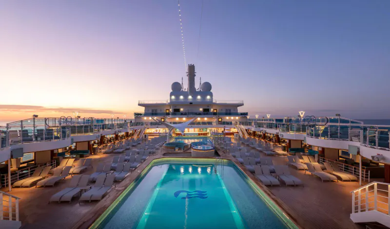 Princess Cruises luxury cruise ship deck with glowing pool featuring 'QC' logo at twilight sunset.
