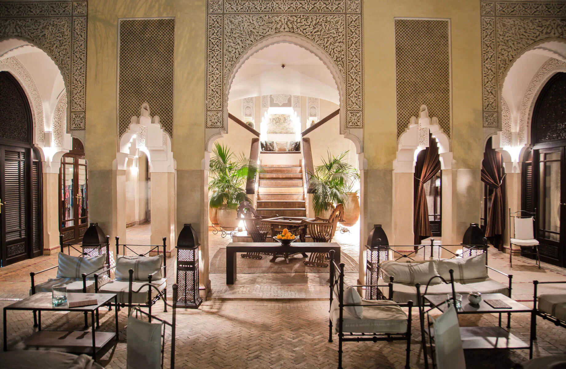 Elegant Moroccan riad courtyard in Marrakech with arched doorways, potted palms, grand staircase, and lantern-lit seating area.