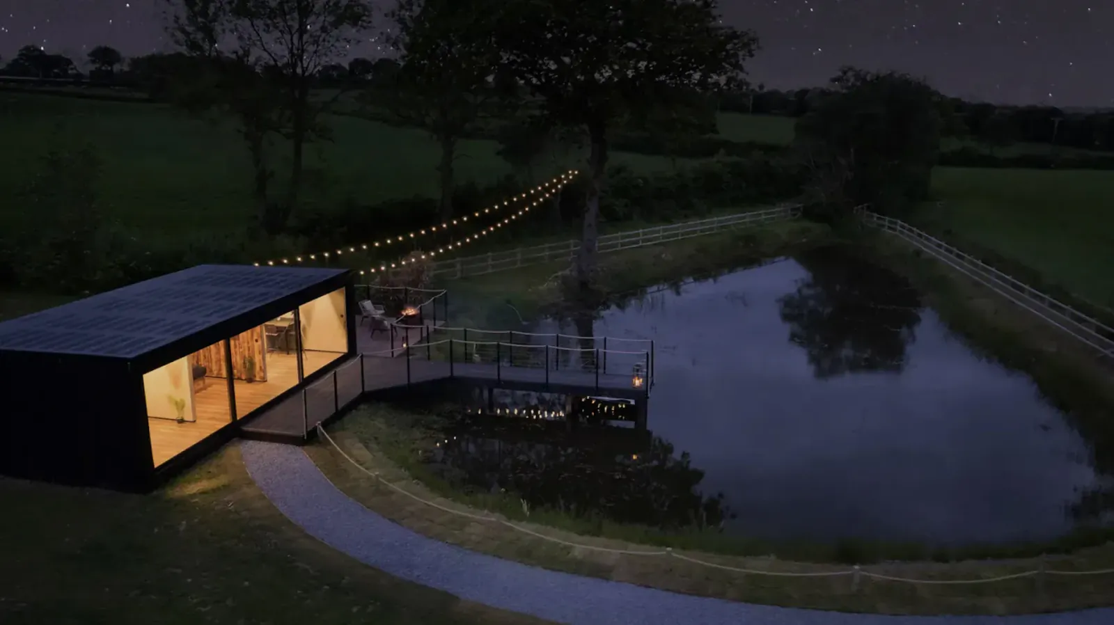 Pantcefn 'The Boathouse' luxury lodge at night by pond, with string lights, bridge, and countryside views.