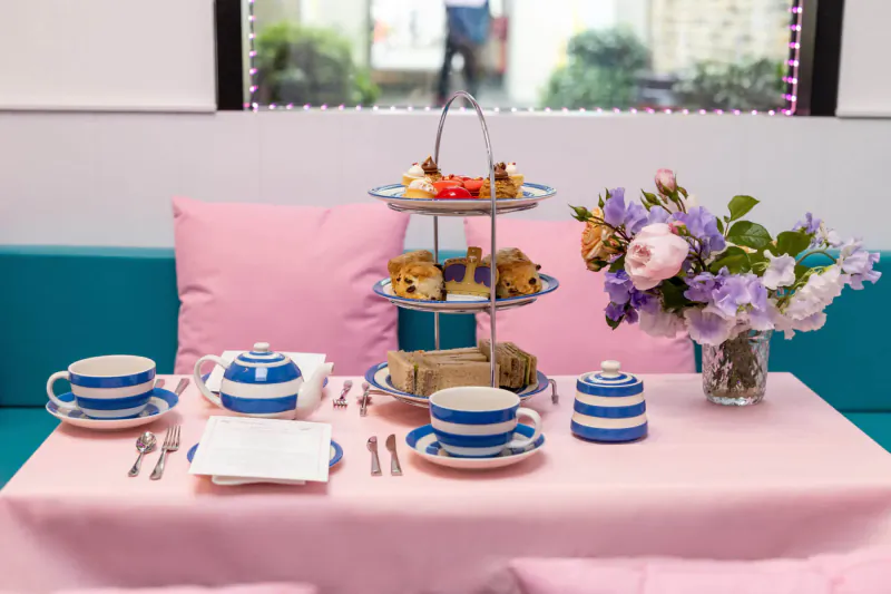 Elegant afternoon tea setup with blue teacups, tiered scones and sandwiches, pink cushions, purple flowers on pink tablecloth by window.