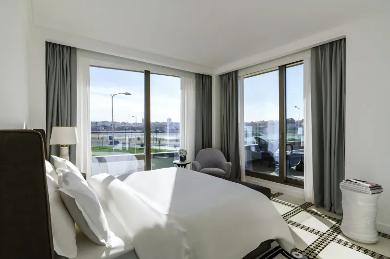 Bright corner hotel bedroom with large windows overlooking marina, white bed, armchairs, and cityscape view.