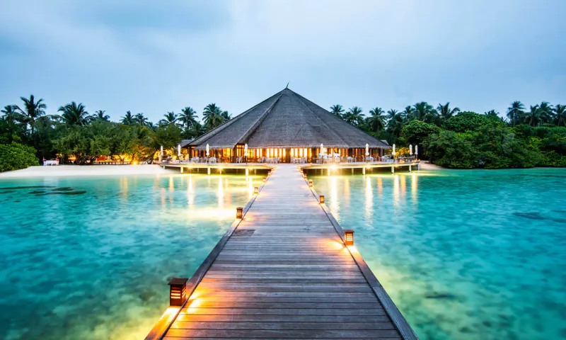 Lit wooden pier leading to thatched roof resort over turquoise lagoon at Hideaway Beach Resort & Spa, twilight.