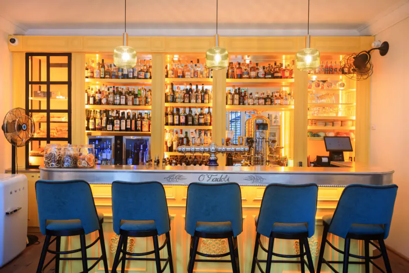 Elegant hotel bar with wooden shelves of liquor bottles, blue stools at curved counter labeled 'Lou Calen', warm pendant lights.