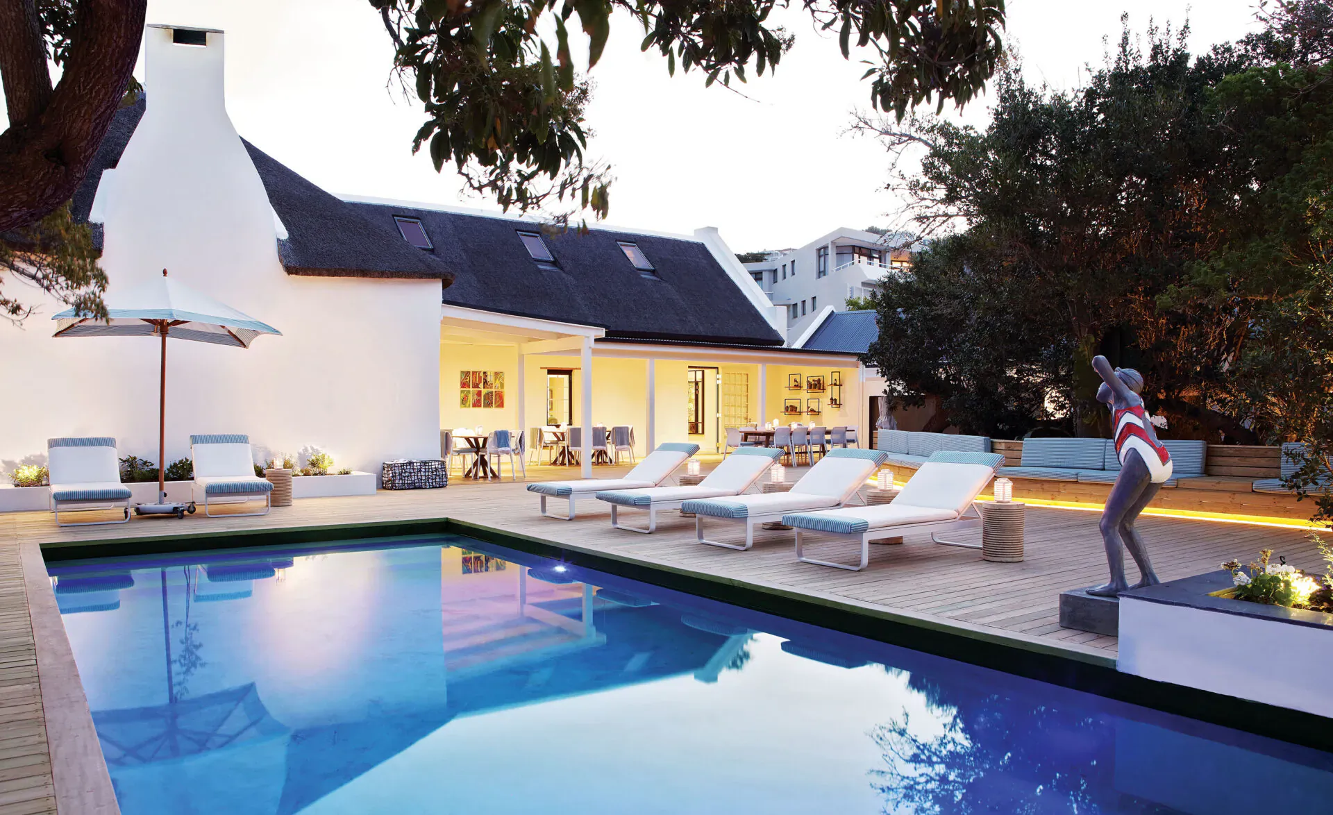 Twilight view of The Old Rectory's blue pool, white lounge chairs, statue, and Cape Dutch building in South Africa
