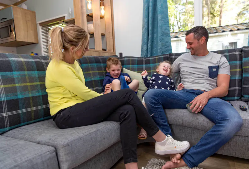 Smiling family of four cuddling and laughing on plaid sofa in cozy cabin kitchen, Sandy Balls Holiday Village.