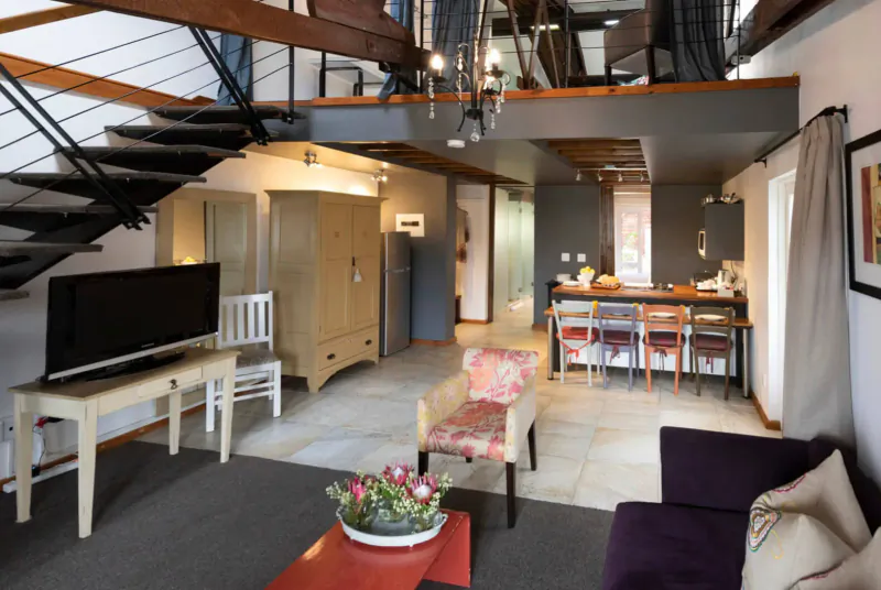 Spacious modern loft interior at The Lofts Boutique Hotel with metal staircase, kitchen, dining area, TV, and plush seating.
