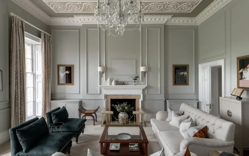 Elegant gray paneled living room in British luxury hotel with chandelier, velvet sofas, marble fireplace, and artwork.
