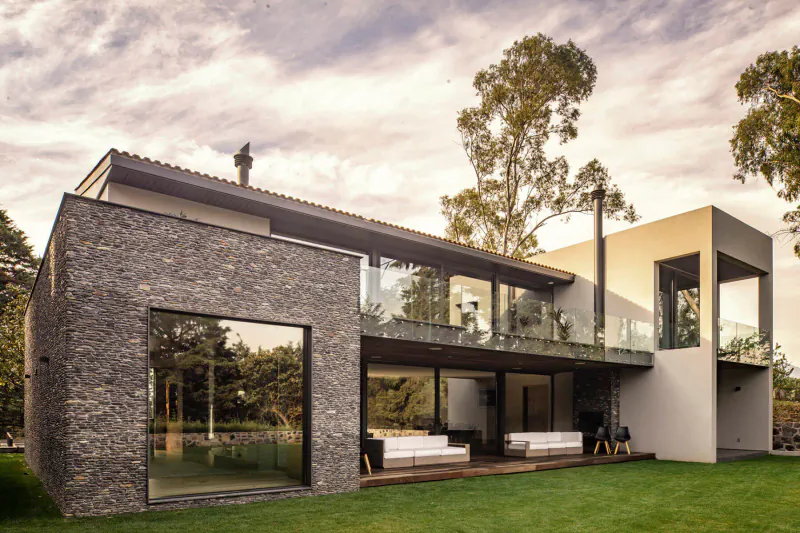 Modern Mexican house with stone and glass facade, expansive balcony with white sofas, lush green lawn, and tall trees at sunset.