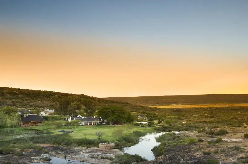 Bushmans Kloof wilderness reserve: luxury retreat with white cottages amid green hills, river, and golden sunset plains.
