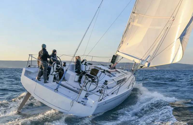 Crew in helmets maneuvers white sailboat speeding through ocean waves at sunset, End of Summer Boat Show.