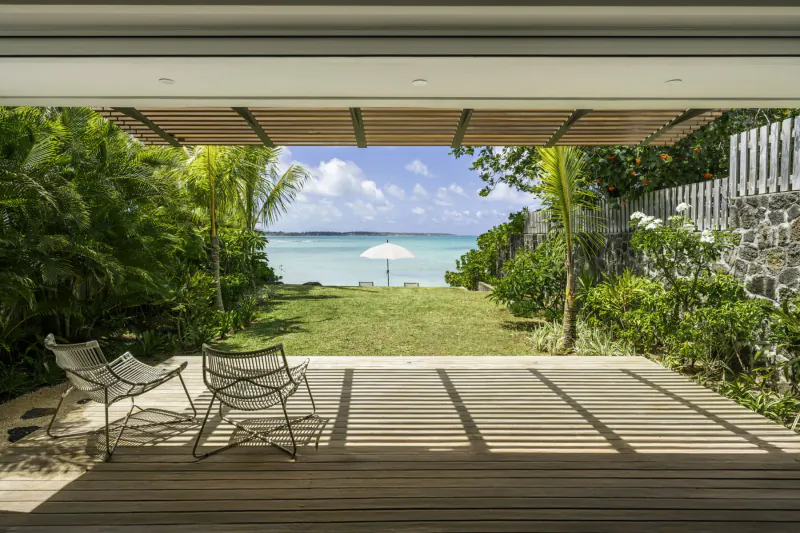 Open wooden deck of Villa Diamond in Mauritius with white chairs overlooking lush garden, lawn, beach, and turquoise sea under blue sky.