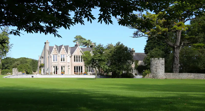 Parc le Breos Country House, a turreted stone mansion with large windows, surrounded by trees and green lawn in Gower, Wales.