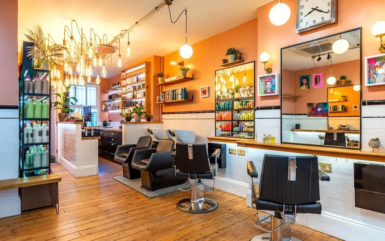Interior of Santi’s London hair salon with black salon chairs, shampoo stations, product shelves, warm pendant lights, and wall clock.