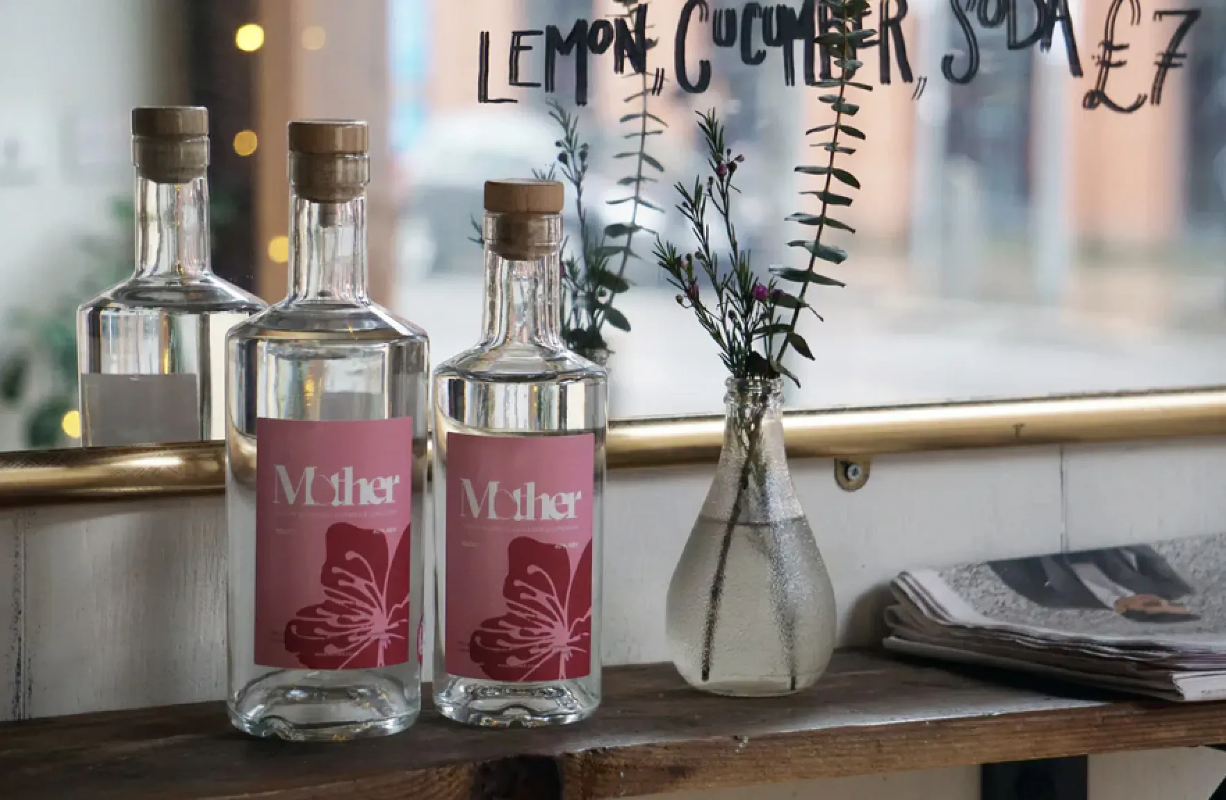 Two Mlk gin bottles with hibiscus labels on wooden shelf beside lemon cucumber spritzer sign, vase of greenery, and newspaper.