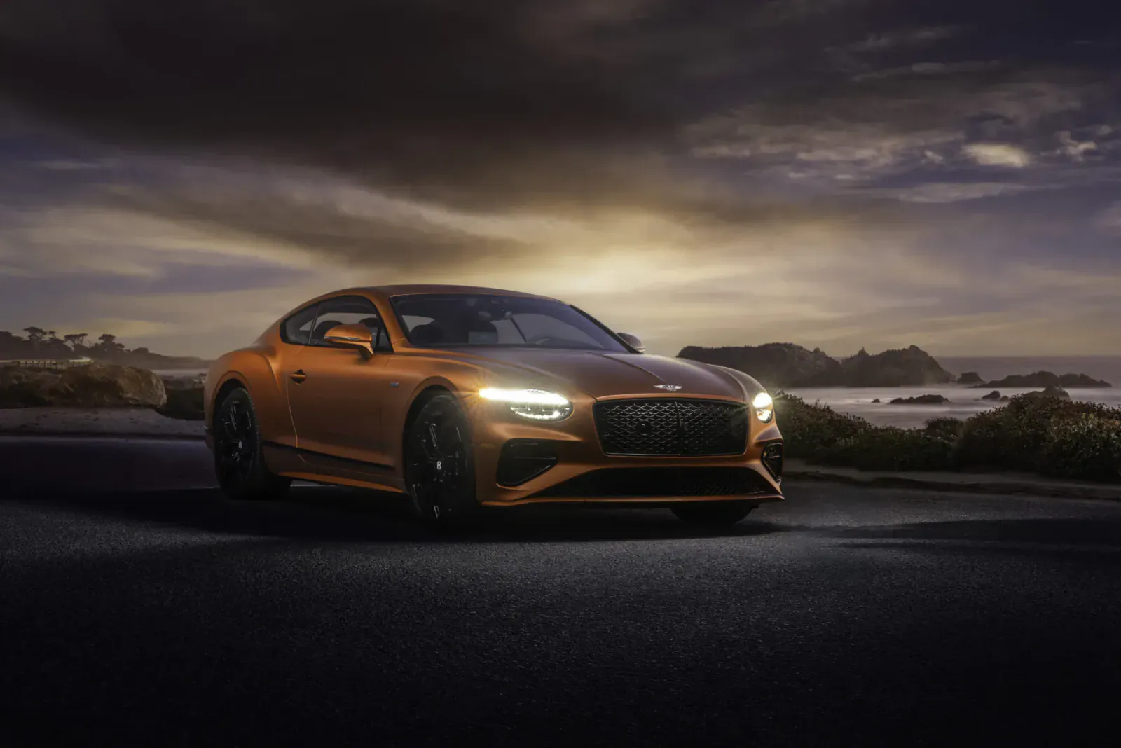 Bentley Continental GT Speed, orange coupe on coastal road at sunset with dramatic sky and ocean.