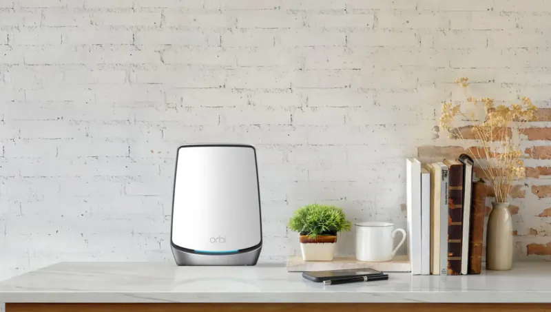 NETGEAR Orbi white WiFi router on wooden table with books, green plant, mug, and phone against brick wall
