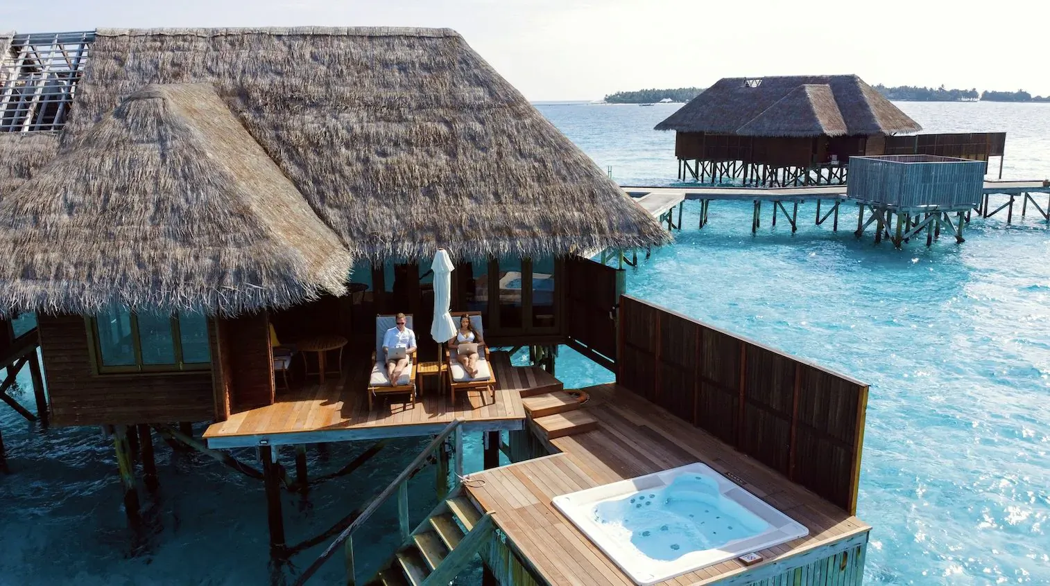 Luxury overwater thatched villa with jacuzzi, lounge chairs under umbrella, and turquoise lagoon