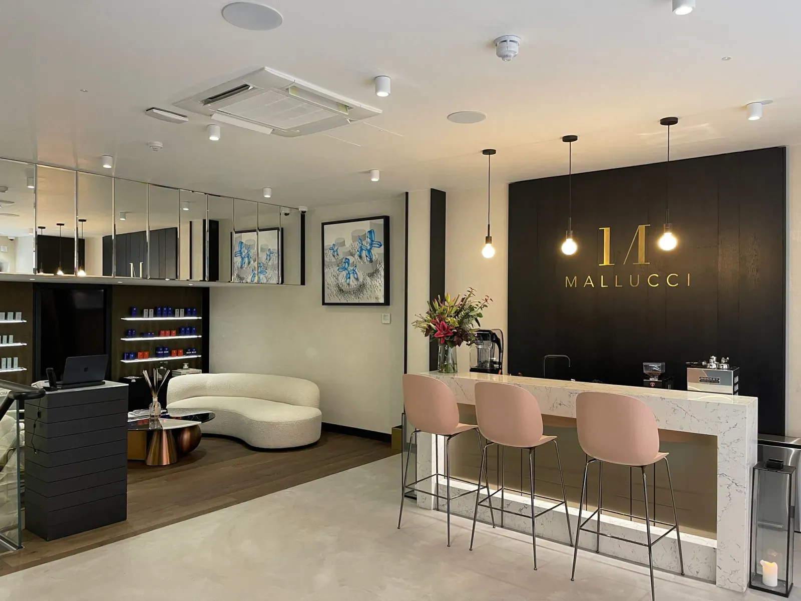 Mallucci London beauty salon reception with white curved sofa, pink stools at white counter, products, and art.