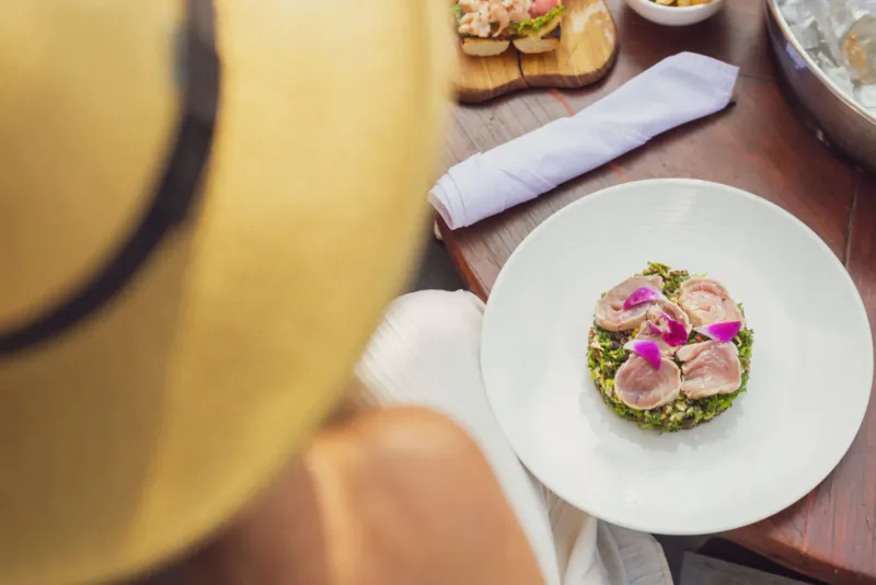 Close-up of tuna tataki on greens with pink flowers on white plate at Catherine’s Café Sur La Plage, Antigua, viewed from above with straw hat brim.