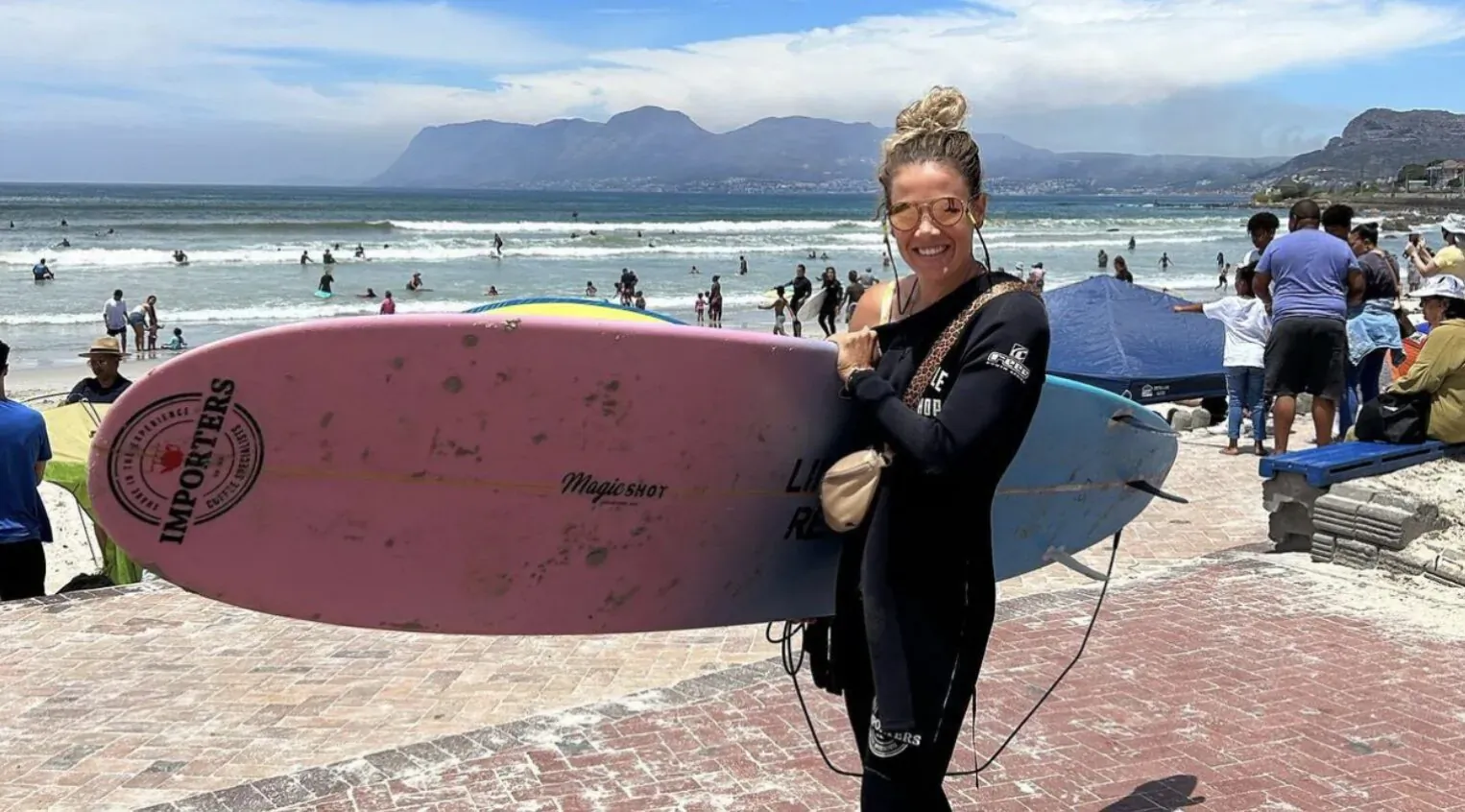 Casey Bennett in black wetsuit holding pink Starboard surfboard on Muizenberg Beach with ocean and mountains behind