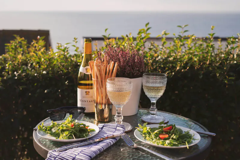Clifftop table with champagne bottle, white wine glasses, breadsticks, salads, and sea view in Cornwall.