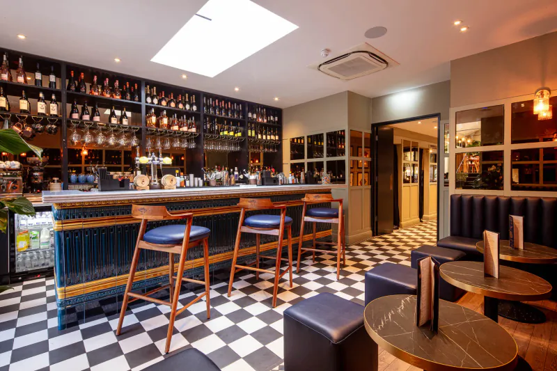 Stylish bar interior at The Mayfair Chippy with wooden shelves of liquor bottles, blue bar stools, checkered floor, and Lobster Pot Booth.