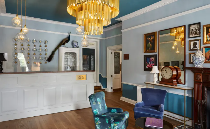 Elegant Hotel Indigo Bath reception with chandelier, peacock decor, vintage clock by fireplace, and blue armchairs.
