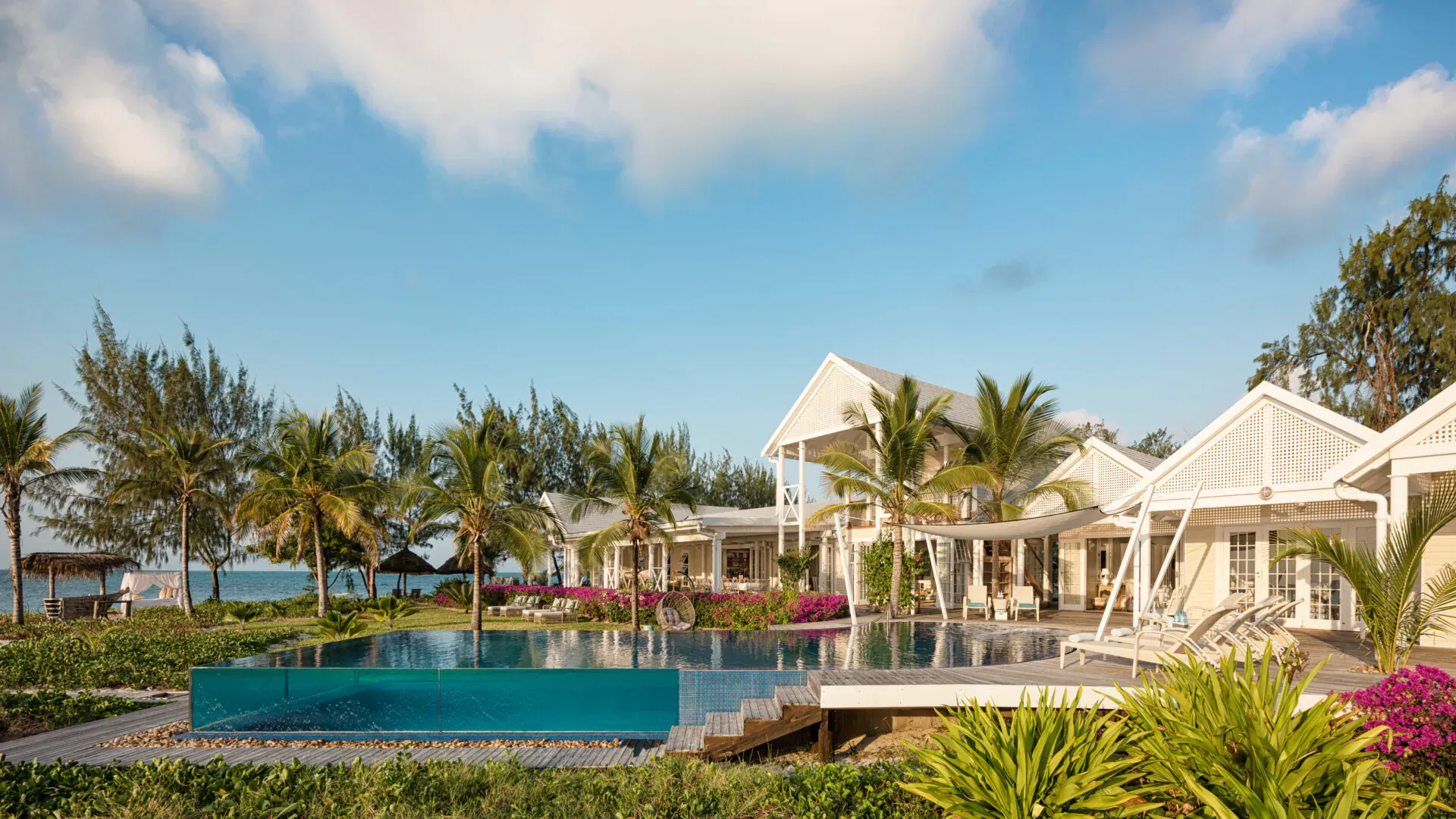 Luxury white villas with infinity pool overlooking beach, surrounded by palm trees and tropical gardens under blue sky