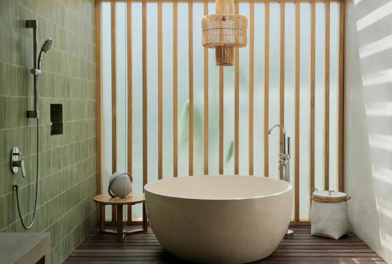 Serene spa bathroom with freestanding beige bathtub, green shower, bamboo screens, rattan lamp, and plants at Amber Lombok Beach Resort.
