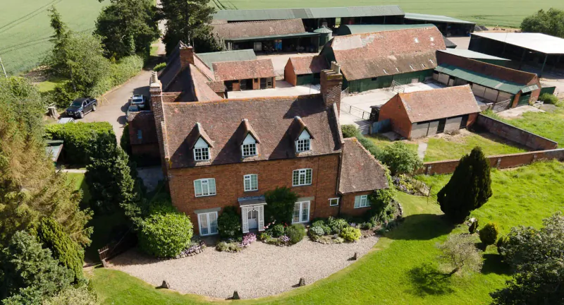 Aerial view of a large red-brick country house with gabled roof, surrounded by barns, trees, and green fields.
