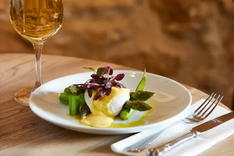 Poached egg with asparagus spears and purple microgreens on sauce, beside white wine glass on wooden table