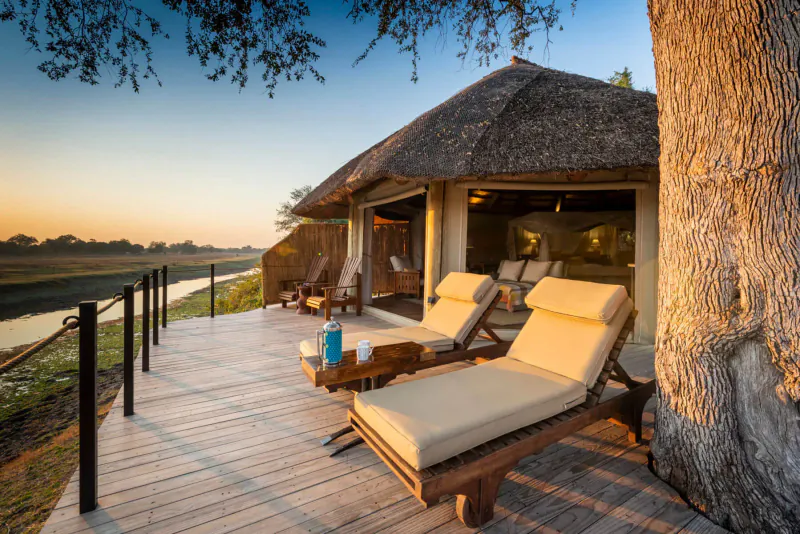 Luxury thatched safari tent with loungers on wooden deck overlooking river at sunset, Lion Camp