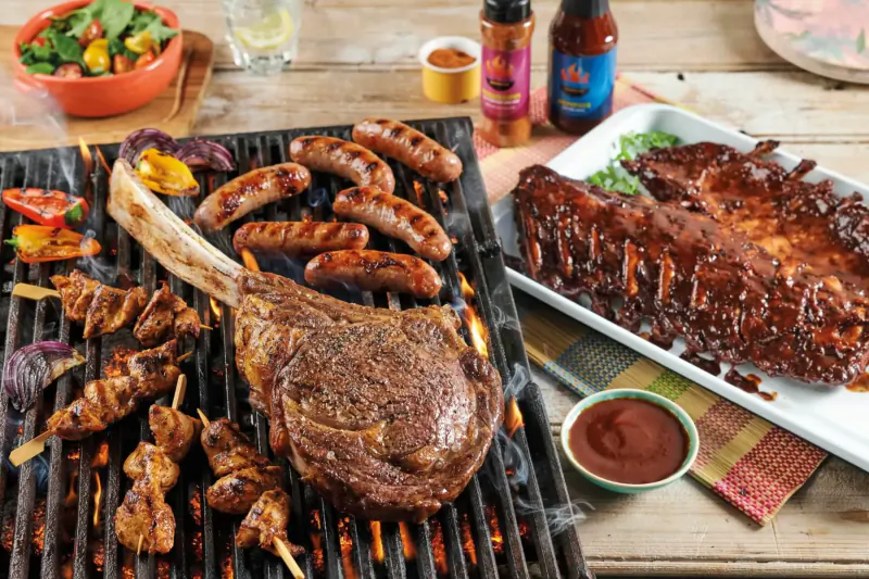 Grilled Tomahawk steak, ribs, sausages, skewers, and peppers on BBQ with sauces and sides.
