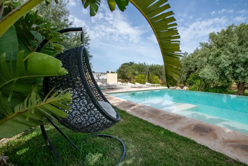 Black rattan hanging egg chair with palm leaves framing a turquoise infinity pool at Zahir Country House in Sicily