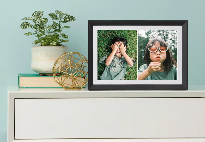 Aura smart photo frame on teal shelf with plant and gold orb, showing two kids: one covering eyes, one blowing with sunglasses.