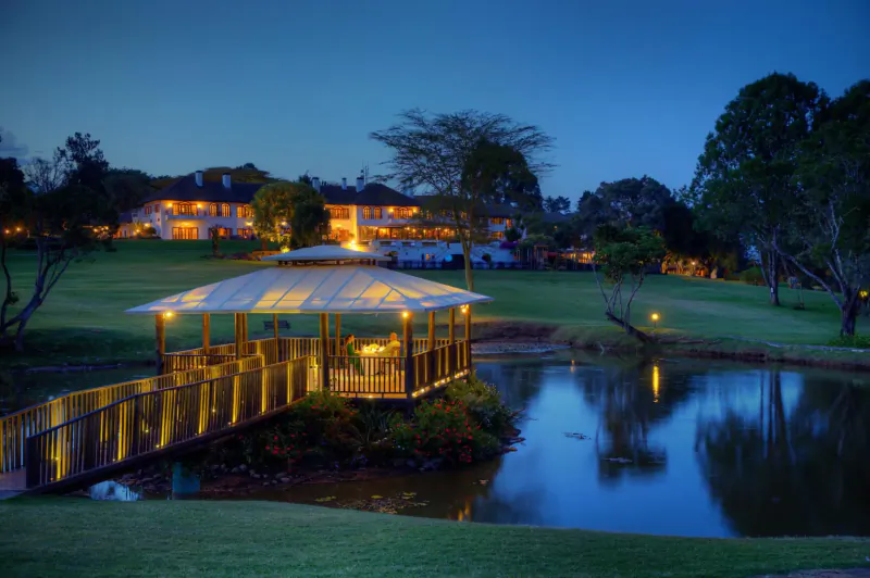 Lit gazebo on island in pond with bridge, surrounded by golf course, trees, and luxury buildings at Mount Kenya Conservancy at dusk