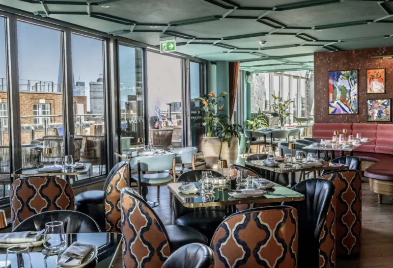 Elegant rooftop restaurant with city skyline view through large windows, ornate teal ceiling, patterned chairs, set tables, and colorful art.