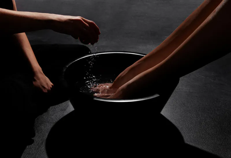 Close-up of hands pouring water into black basin for foot soak in dim-lit spa setting