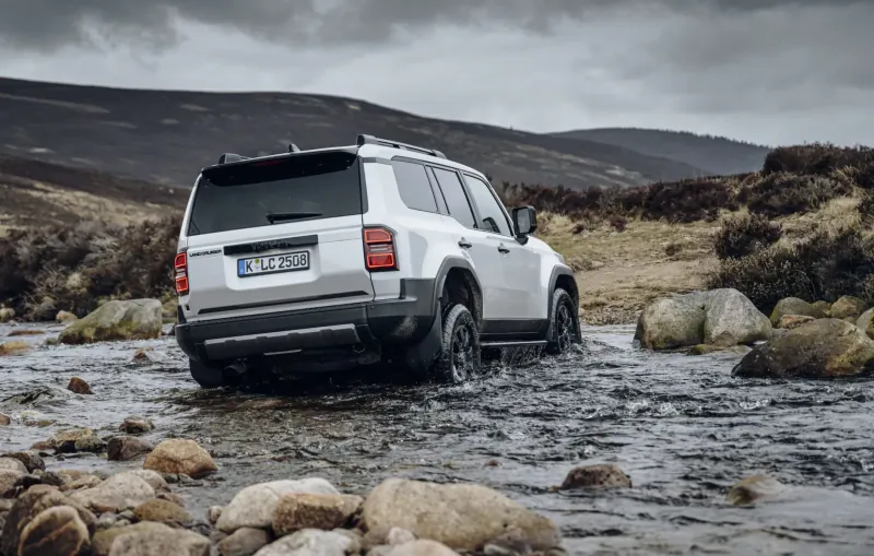 White Toyota Land Cruiser SUV driving through shallow stream in rugged Scottish hills, rear view.