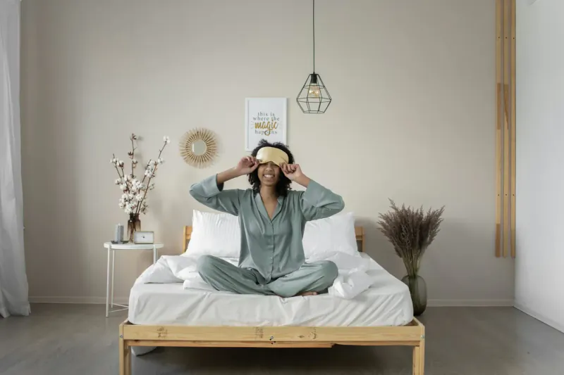 Smiling Black woman in sage green pajamas sits cross-legged on bed, wearing eye mask, in elegant bedroom with plants and pendant light.