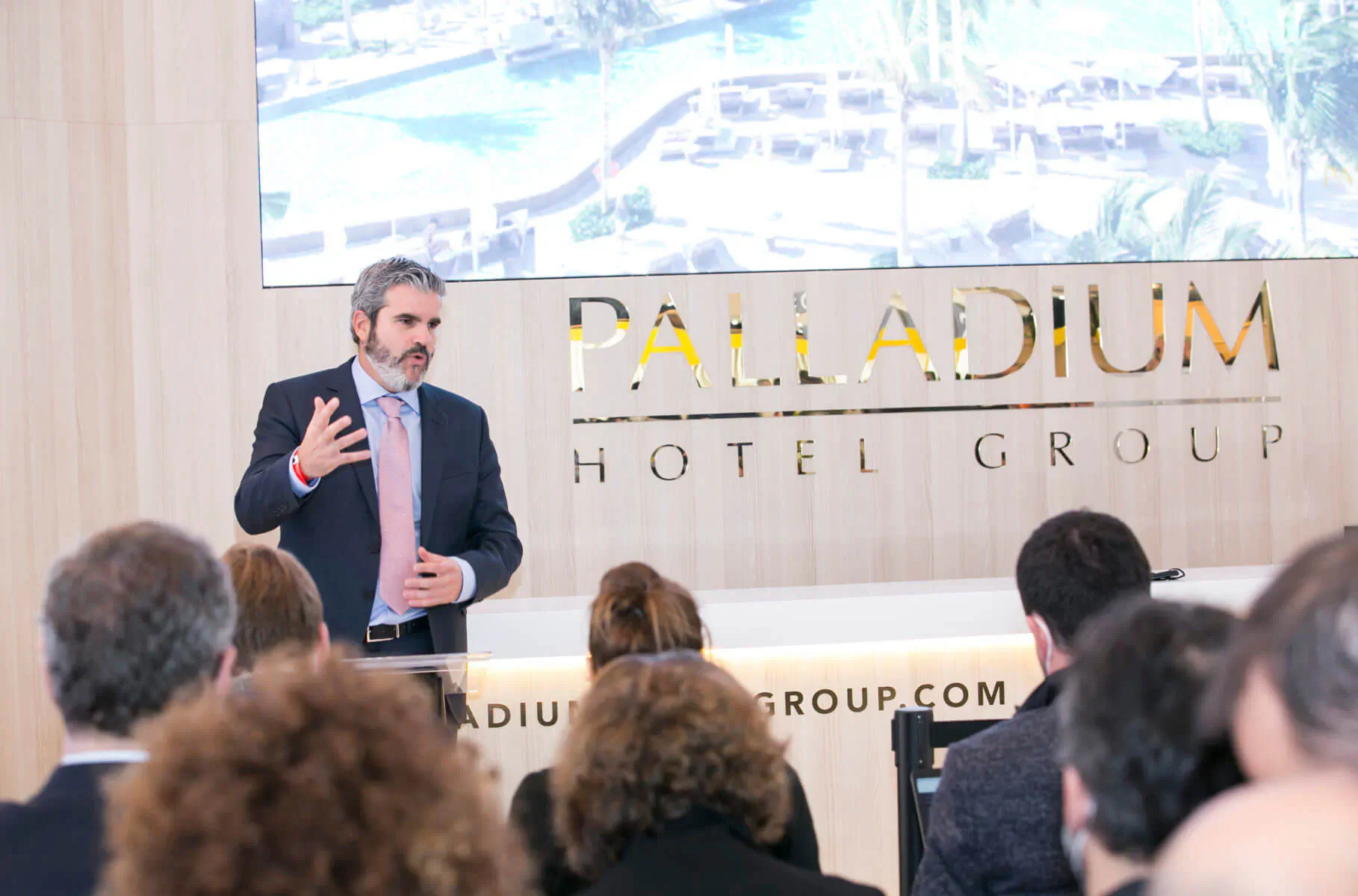 Man in suit presenting at Palladium Hotel Group event with audience and screen, backdrop reads 'Palladium Hotel Group'.