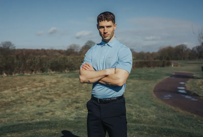 Man in light blue polo and black trousers stands arms crossed on sunny golf course green.
