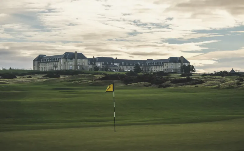 Fairmont St Andrews hotel atop grassy dunes with golf flag on green course under cloudy sky, Scotland.