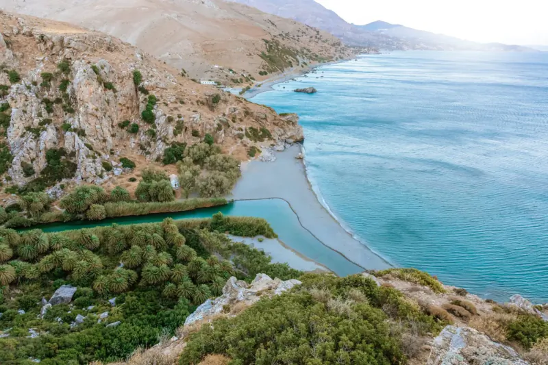 Aerial view of Candia Park Village in Crete: rugged hills, green river, sandy beach, and turquoise sea.