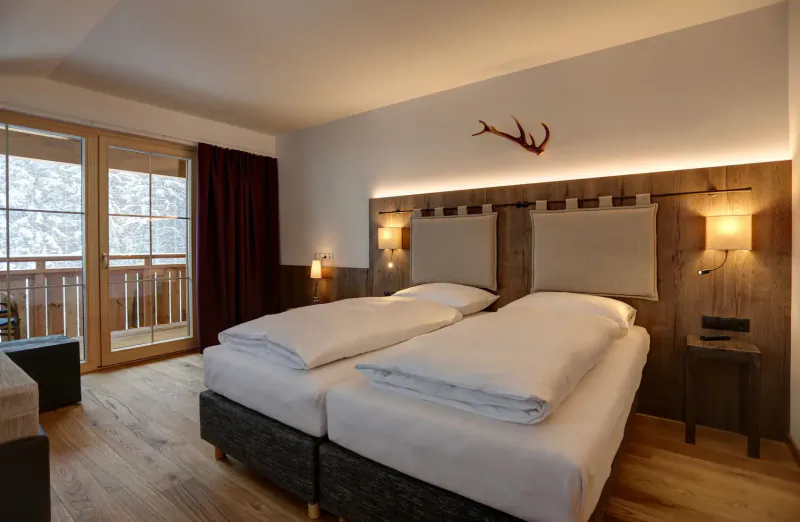 Cozy Hotel Kösslerhof room with two twin beds, antler decor, balcony view of snowy mountains.